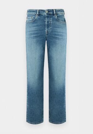 Kvinde iført lyseblå denimjakke, matchende jeans, hvid top, hoops-øreringe og sorte sandaletter med hæl mod hvid baggrund.