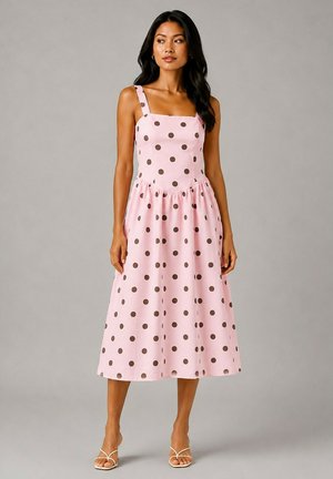 Femme aux longs cheveux foncés porte une robe midi rose sans manches avec des pois marron et des talons blancs à brides, debout devant un fond uni.