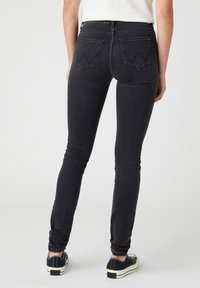 Jeans ajustados negros de tiro alto, con dos bolsillos traseros que presentan una costura distintiva en forma de Y, combinados con zapatillas negras y una camiseta sencilla.