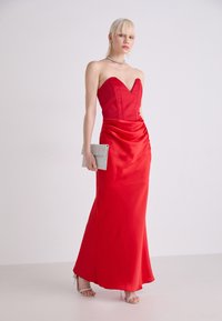 Rochie din satin roșu cu un gât în formă de inimă, corset ajustat și un detaliu drapat la talie. Asortată cu o poșetă argintie și sandale cu cureluțe.