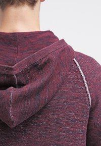 Mörkröd texturerad huvtröja med en åtsittande design och grå sömmar, med en dragsko i huvan och ribbade detaljer. Synliga tygdetaljer.