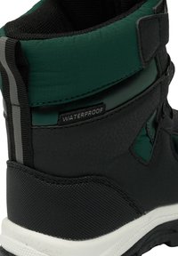 Musta ja vihreä vedenpitävä saapas, jossa on teksturoitu musta alaosa, vihreä kangasyläosa ja heijastavia harmaita yksityiskohtia.