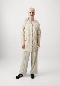 Beige vanntett jakke med hette, trykknapper og to frontlommer, sammen med vide, lyse bukser og svarte sneakers.