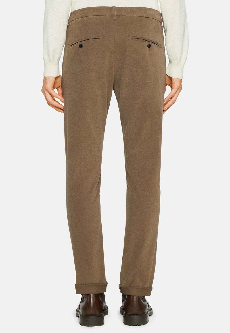 Pantalons marron sur mesure avec une texture douce, présentant deux poches arrière avec des fermetures à bouton et un ourlet roulé, associés à des chaussures foncées.