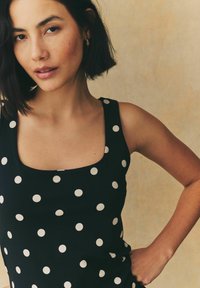 Black sleeveless top with a square neckline and white polka dots, smooth fabric, fitted shape, minimalistic design.