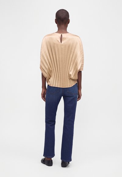 Personne de dos portant un haut beige plissé avec un détail dos en forme de serrure, un jean bleu droit et des chaussures noires.
