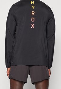 Camisa atlética de manga larga negra con letras multicolores "HYROX" impresas verticalmente en la parte posterior. La tela parece suave y ligera.