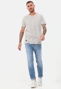 Jasnoszara koszulka z krótkim rękawem, okrągłym dekoltem i kieszonką na piersi, zestawiona z jasnoniebieskimi dżinsami i białymi trampkami. Prosty, casualowy design.