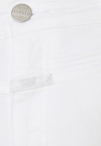 Denim blanco con un primer plano de un botón plateado con la inscripción "CLOSED" grabada. Incluye una etiqueta de tela y detalles de costura visibles.