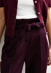 Cardigan bordeaux avec des boutons dorés sur un débardeur blanc à côtes. Pantalons bordeaux taille haute avec une ceinture à nouer et des poches latérales.