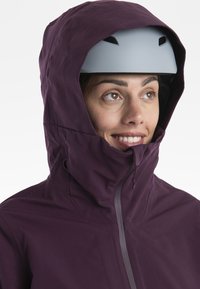 Fioletowa wodoodporna kurtka z zamkiem błyskawicznym z przodu i regulowanym kapturem, noszona z jasnoszarym hełmem, podkreślająca gładką tkaninę i funkcjonalny design.