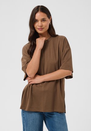 Femme aux longs cheveux foncés portant un t-shirt marron ample et un jean bleu, posant avec une main près du visage et l'autre croisée sur la taille.