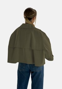 Olivgrön kort jacka med bred krage, lager-på-lager design på baksidan och långa ärmar. Bärs över höga blå jeans.