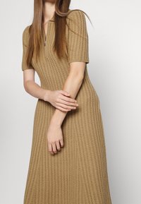 Strikket kjole i lys brun med et tekstureret, ribbet design, korte ærmer og krave med lynlås. Figursyet silhuet med en udvidet kant.