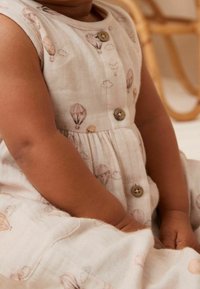 Småbarn iført en beige kjole med vintage luftballonprints og træknapper, siddende indendørs på en lys overflade.