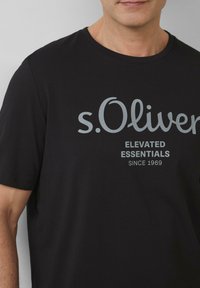 Czarny bawełniany t-shirt z okrągłym dekoltem i krótkimi rękawami, ozdobiony szarym logo oraz napisem "s.Oliver Elevated Essentials."