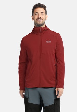 Mann trägt eine rote Jack Wolfskin Zip-Up-Jacke und graue sowie schwarze Outdoor-Hosen, lächelnd und vor einem einfarbigen Hintergrund stehend.