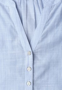 Chemise bleu clair à boutons avec un subtil motif de fines rayures verticales et un col en V. Trois boutons blancs ferment le devant.