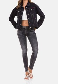 Schwarze Jeansjacke mit weißer Naht, Fronttaschen und Knopfverschluss. Ein Paar ausgebleichte schwarze Skinny-Jeans mit glatter Oberfläche. Weiße Sandalen mit Riemen.