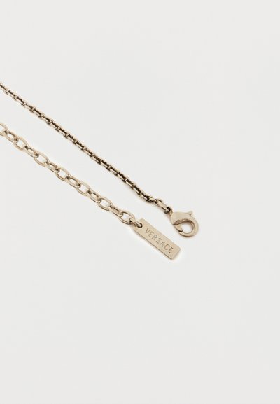 Αλυσίδα σε χρυσό τόνο με μια μικρή ορθογώνια ετικέτα χαραγμένη με τη λέξη "VERSACE" και κούμπωμα σε σχήμα αστακού σε λευκό φόντο.