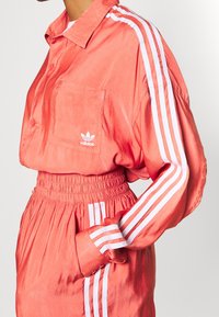 Korallin värinen urheiluasu, joka sisältää napillisen paidan, jossa on tasku ja valkoinen Adidas-logo, yhdistettynä kuminauhavyötäröisiin housuihin, joissa on raidallisia yksityiskohtia.