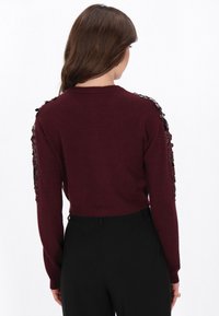 Burgunderfarbener Strickpullover mit strukturierten Blumenapplikationen an den Ärmeln, geripptem Saum und rundem Halsausschnitt, kombiniert mit schwarzen schmalen Hosen.