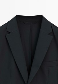 Blazer nero con revers a punta, due tasche anteriori e superficie liscia. L'interno è semplice e con una finitura pulita.