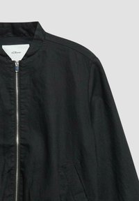 Veste noire zippée avec col montant, poche latérale et fermeture éclair argentée visible, présentée sur un fond gris clair uni.