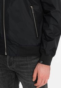 Veste bomber noire avec un extérieur en nylon brillant, des poignets côtelés, une poche zippée sur le côté et une taille avec un détail d'étiquette. Surface texturée.