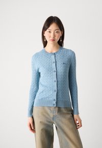 Light blue knitted cardigan with cable pattern, round neckline, and button closure. Ribbed cuffs and hem, paired with beige jeans.