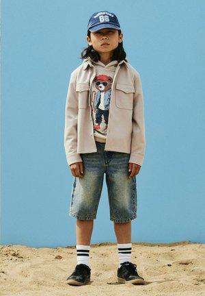 Lapsi seisoo hiekalla sininen lippalakki päässään, beige takki, karhukuosinen huppari, farkkushortsit, valkoiset raidalliset sukat ja mustat lenkkarit päällään sinistä taivasta vasten.