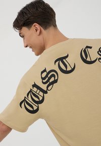 T-shirt in cotone beige con grande lettering gotico nero sulla schiena, maniche corte e design a vestibilità ampia.
