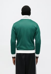 Green athletic sweatshirt with a high collar, white trim at the cuffs and hem, smooth texture, and a relaxed fit, shown from the back.