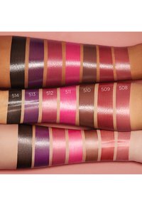Échantillons de produits pour les lèvres sur les bras, présentant diverses teintes : brun foncé, violet, rose et nude. Les finitions sont brillantes, avec des numéros visibles au-dessus de chaque échantillon.