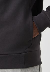 Felpa grigia scura con una texture liscia, polsini a coste, tasche laterali e un girovita aderente. Il taglio mette in mostra un design rilassato e moderno.