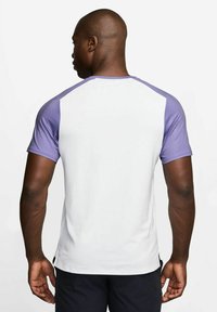Kortermet treningsskjorte med hvit kropp og lilla raglan-ermer, laget av et glatt, lett stoff. Baksiden viser et enkelt design.