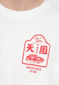 Bílé tričko s červenou grafickou nálepkou s japonským textem a stylizovanou ilustrací vepřového řízku. Jednoduchý, bavlněný materiál.