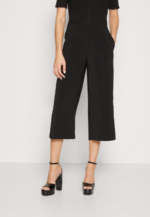 Femme portant un pantalon noir large court et des talons hauts noirs avec des brides à la cheville, debout devant un fond clair uni.