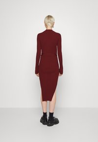 Personne avec des cheveux blonds courts portant une robe midi bordeaux à côtes ajustée avec une fente latérale et des chaussures noires épaisses, se tenant contre un mur blanc.