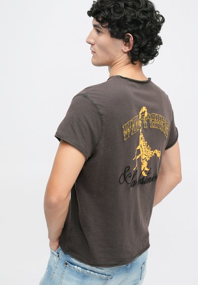 T-shirt de algodão castanho com decote redondo e mangas curtas. Apresenta impressão gráfica amarela e texto preto nas costas, com bainha desfeita.