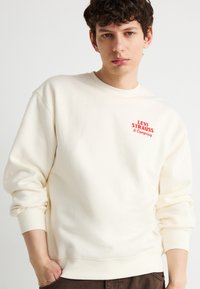 Νέος άντρας που φοράει κρεμ φούτερ Levi's με κόκκινο λογότυπο, ένα χέρι στην τσέπη, μπροστά σε απλό ανοιχτό φόντο.