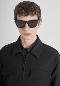 Occhiali da sole neri con montatura angolare, lenti scure, indossati con una giacca nera a zip con colletto e due tasche frontali.