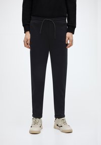 Μαύροι φόρμες με ελαστική μέση και κορδόνι, με εφαρμοστή γραμμή και μανσέτες στους αστραγάλους, συνδυασμένες με μπεζ αθλητικά παπούτσια.