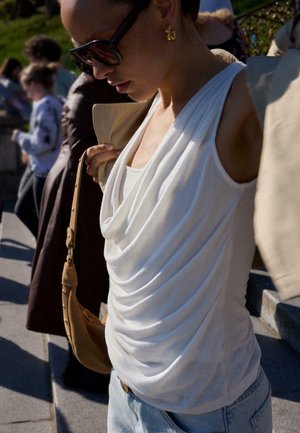 Žena s slunečními brýlemi a zlatými kruhovými náušnicemi si venku upravuje béžový kabát přes bílý bezrukávový top s volánem a světle modré džíny.