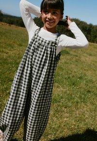 Jeune fille souriante en plein air, ajustant ses cheveux, portant un haut blanc à manches longues et une salopette à carreaux noir et blanc dans un champ herbeux.