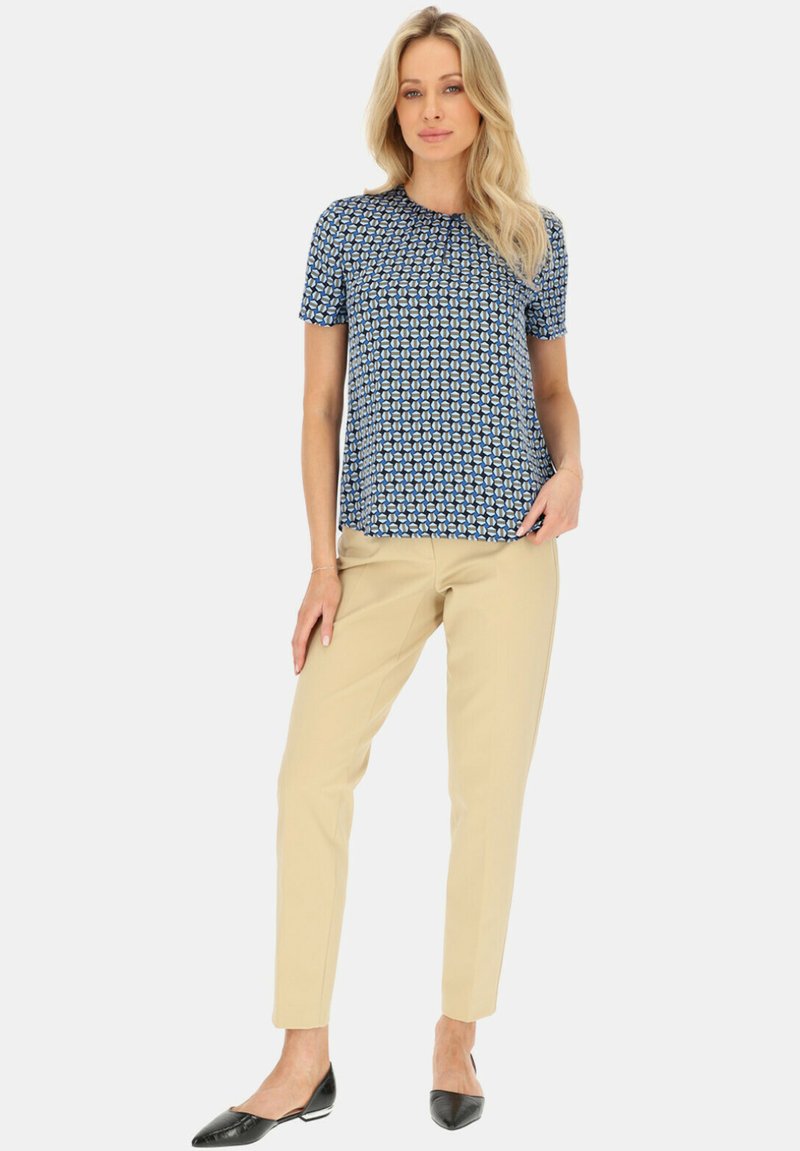 Niebieska bluzka z krótkim rękawem w geometryczny wzór, w połączeniu z beżowymi spodniami typu tapered i czarnymi balerinami. Gładki materiał o luźnym kroju.