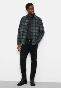 Mann steht in einer blau-grünen karierten Jacke über einem dunklen Hemd mit Knöpfen, schwarzen Hosen und schwarzen Schnürstiefeln vor einem weißen Hintergrund.