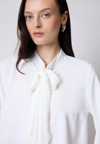 Blusa blanca con una corbata plisada y fluida en el cuello. La tela es suave, con un diseño minimalista y sin patrones ni adornos visibles.