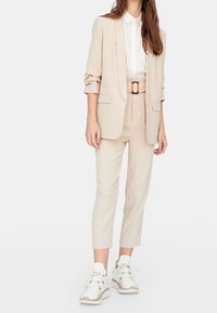 Béžový upravený blazer s ohrnutými rukávy přes bílou košili, v kombinaci s béžovými kalhotami s vysokým pasem a bílými teniskami s vzorem.