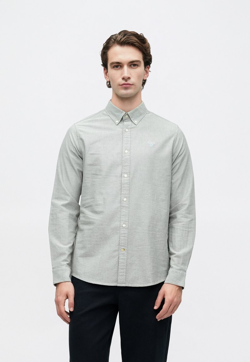 Jovem com cabelo curto e encaracolado, vestindo uma camisa abotoada cinza-claro e calças escuras, em pé contra um fundo branco simples.